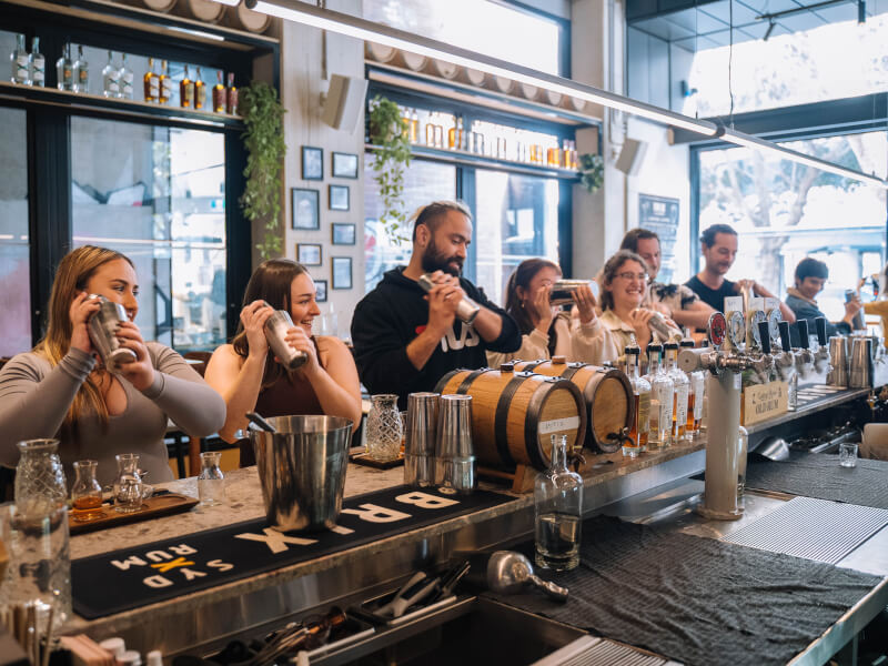 A line of people smiling and shaking cocktails in front of a bar. 